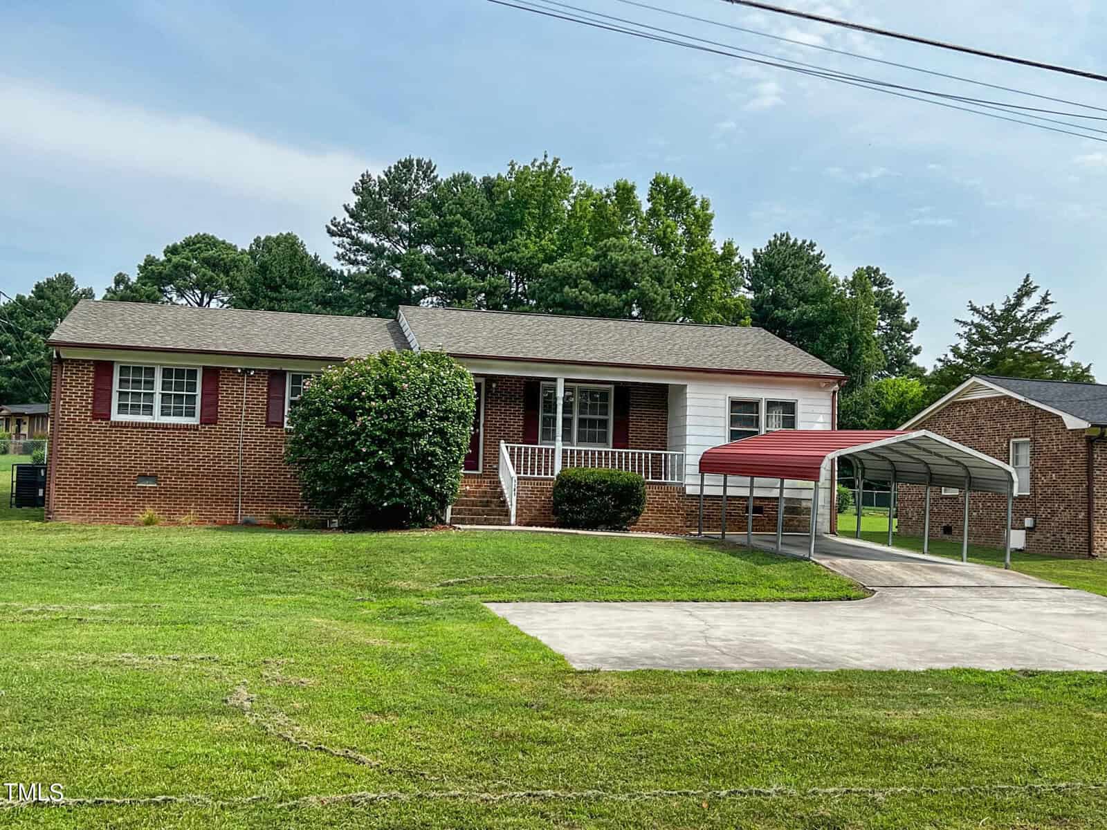 A single-story brick house with a gray roof and white trim. It has a small front porch with white railings, a manicured lawn, and a driveway leading to a carport with a red roof. Tall trees are in the background.