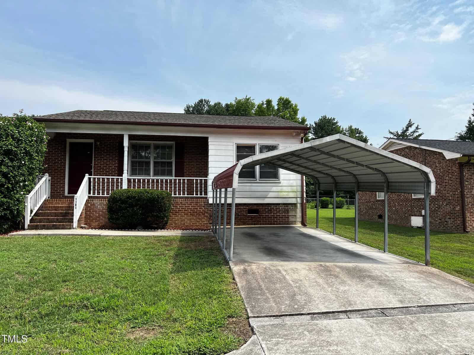 A single-story brick house with a white front facade and front steps leading to the entrance. A large, metal carport is positioned over the driveway. The house has a small front yard with green grass and a hedge near the steps. The sky is clear with few clouds.