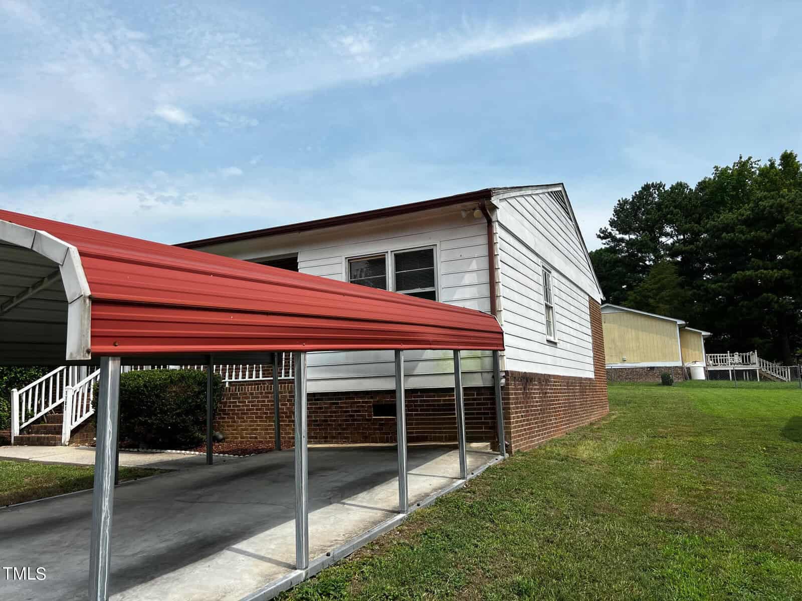 A one-story white house with a brick foundation has a red metal carport attached. The carport covers a concrete driveway. The house has several windows, and there's a grassy yard on the right side. Trees and a second building are visible in the background.
