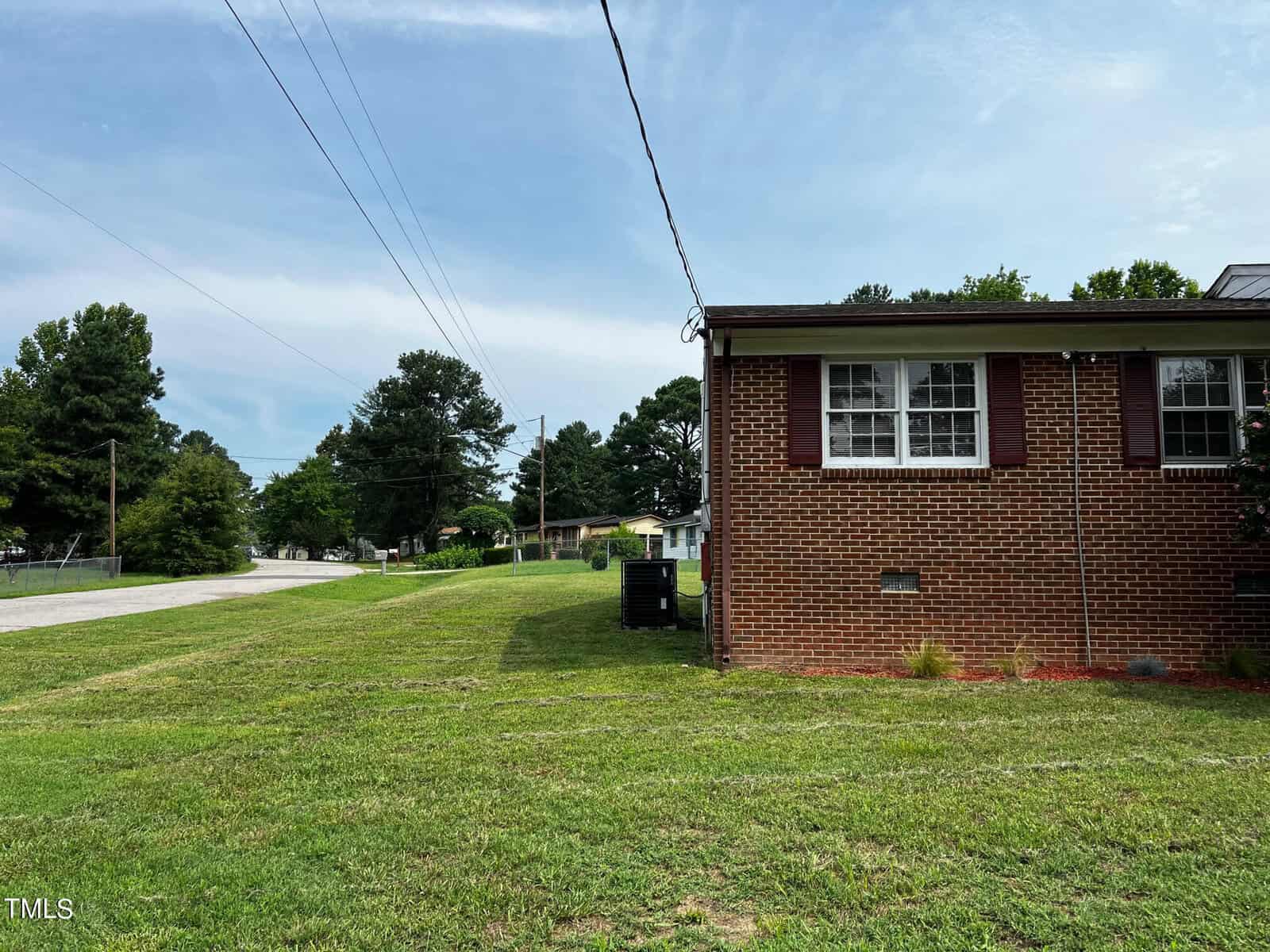 A brick house with a grassy front yard on a clear day. The house features white-framed windows with shutters. Overhead power lines lead down the street, which is bordered by trees and greenery. The sky is partly cloudy, adding to the serene suburban setting.