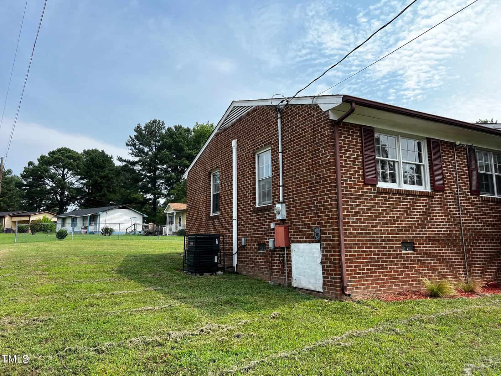 A brick house with white trim and multiple windows is situated on a neatly cut grassy lawn. An air conditioning unit is attached to the side of the house. The sky is partly cloudy, and neighboring houses are visible in the background along with some trees.