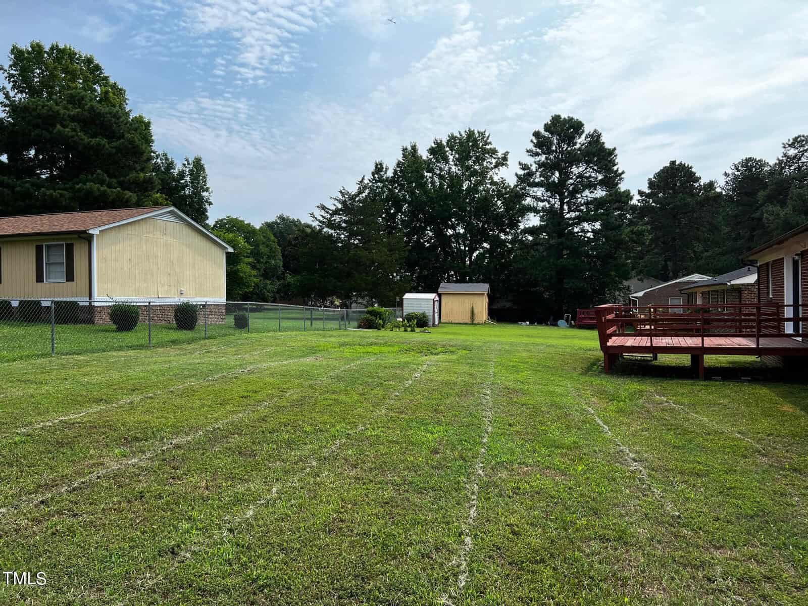 A spacious, freshly cut grassy backyard with a wooden deck on the right side. There are a few houses in the background, including a yellow building on the left with a chain-link fence and another wooden structure toward the center. Trees line the background.