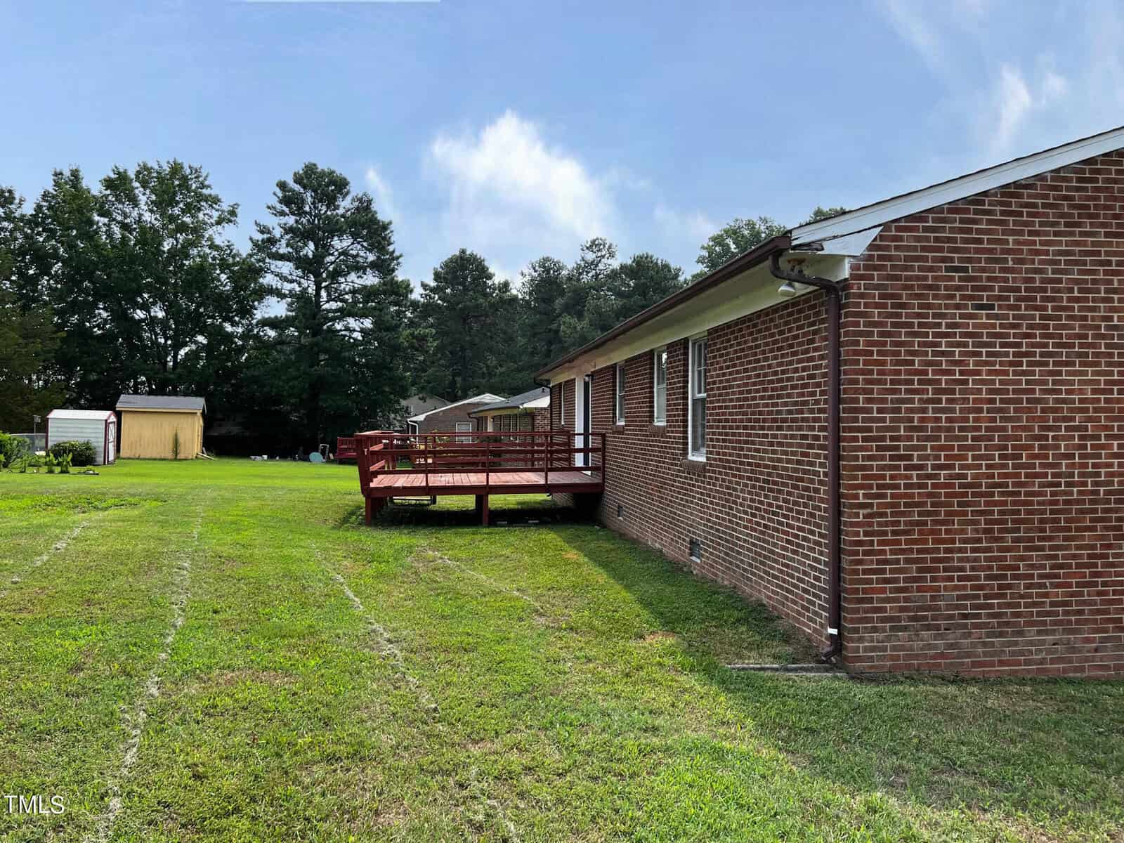 A single-story red brick house with a spacious backyard and a wooden deck attached. The yard is large and grassy, with a yellow shed in the distance. Trees line the edge of the property, and a clear sky is visible.