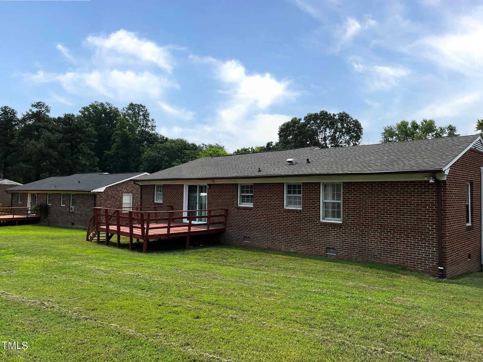 A single-story brick house with a dark roof is shown from the backyard. There is a wooden deck attached to the house. The yard has well-maintained grass and is bordered by trees in the background. Other similar houses are visible in the distance.