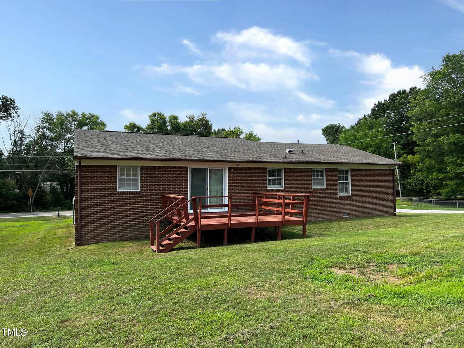 A single-story brick house with a dark roof and a large wooden deck at the back. The house has sliding glass doors opening onto the deck and is surrounded by a well-maintained grassy yard. The background has trees and a partly cloudy sky.