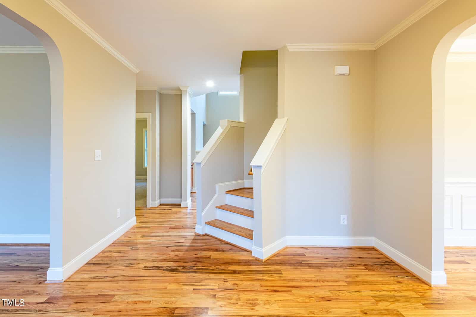 Interior view of a home featuring a staircase with wooden steps and white railings against a beige wall. The room has hardwood floors and archways leading to adjoining spaces. Natural light illuminates the area.
