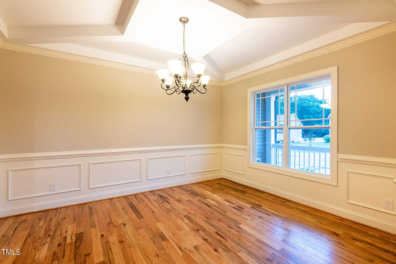 A bright dining room with hardwood floors, a decorative ceiling, and an elegant chandelier. The walls feature wainscoting, and a large window provides natural light, offering a view of a porch and greenery outside.