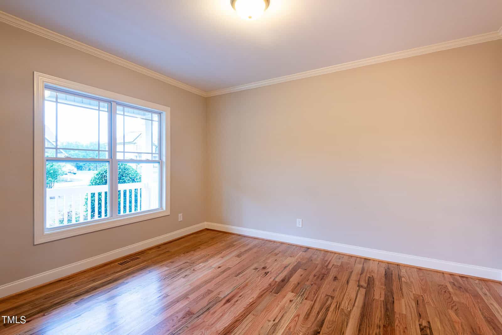 An empty room with beige walls and wooden floors, featuring a large window with white trim. Natural light enters through the window, and a simple ceiling light fixture is present.