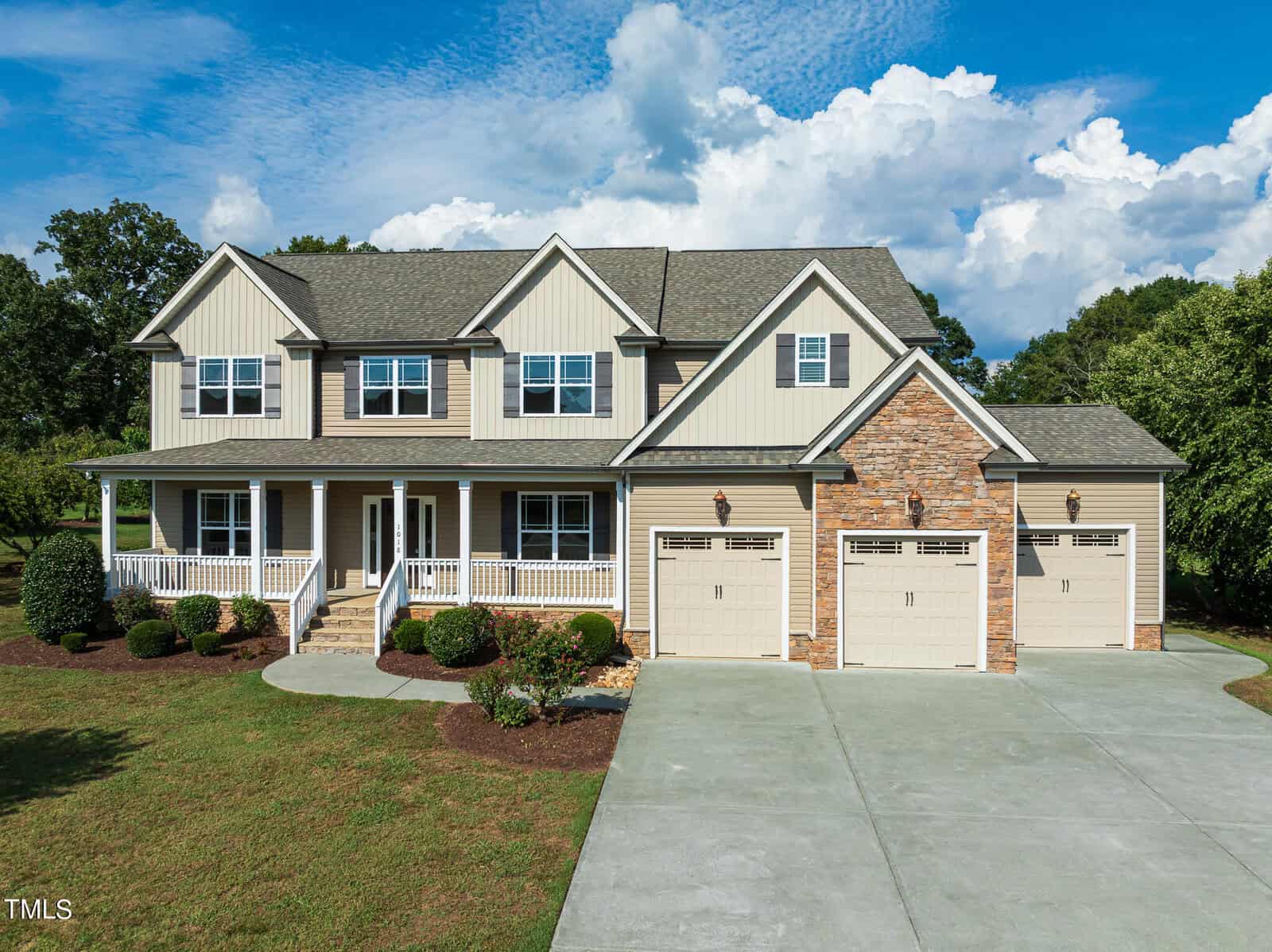 A large two-story house on 1018 Bluebell Lane, with beige siding and stone accents, features a welcoming front porch, three garage doors, and a neatly manicured lawn. Set in the charming Wake Forest area, the scene is framed by a blue sky with scattered clouds.