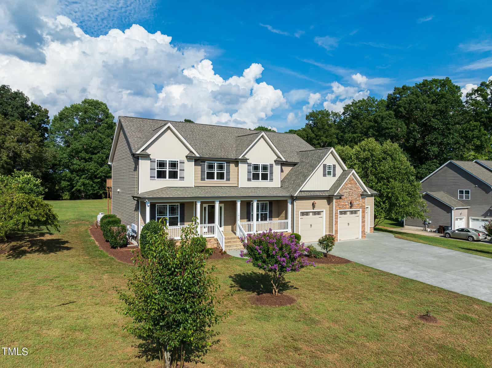 A large two-story house with beige siding and stone accents, featuring a front porch and three-car garage. It's set in a spacious yard with well-maintained grass, a few trees, and surrounded by greenery under a blue sky with clouds.