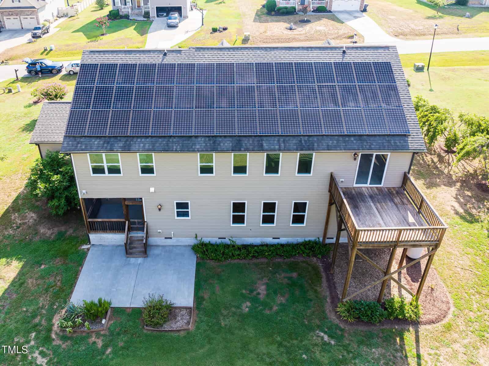 Aerial view of a two-story house with rooftop solar panels. The house has a wooden deck, multiple windows, and is surrounded by a grassy yard. Other homes and streets are visible in the background.