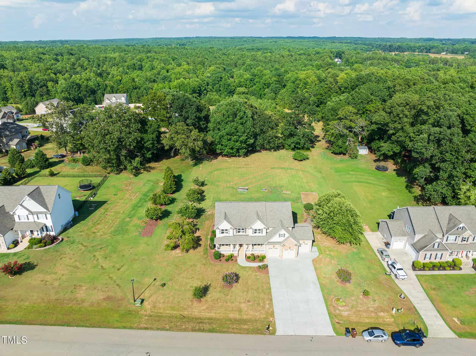 Aerial view of a suburban neighborhood with several houses surrounded by lush green trees and lawns. The homes have driveways, and a few cars are parked outside. The sky is partly cloudy, enhancing the vibrant greenery of the landscape.