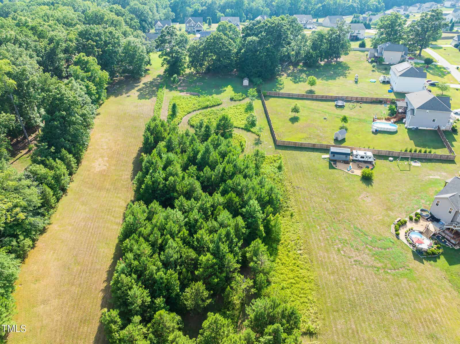 Aerial view of a rural area featuring green fields and clusters of trees. There are several houses with fenced yards and scattered agricultural plots. The landscape is surrounded by dense forest and residential properties.