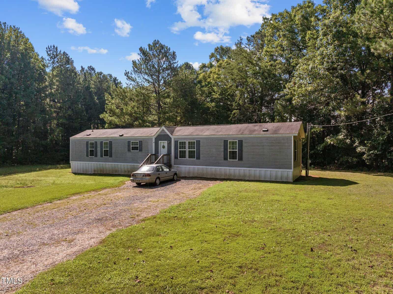 Located at 4752 Old Nc 75, this single-story mobile home in Oxford features a gray exterior and a quaint porch on a grassy lot. A gravel driveway guides you to a parked car, all framed by tall trees under a partly cloudy sky.