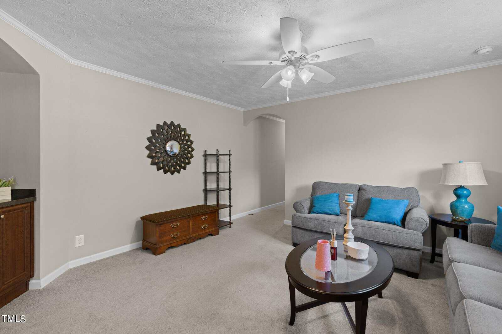 A cozy living room with light gray walls and a ceiling fan. There's a gray couch with blue pillows, a round coffee table with candles, a decorative mirror on the wall, a wooden chest, and a black metal shelf in the corner.
