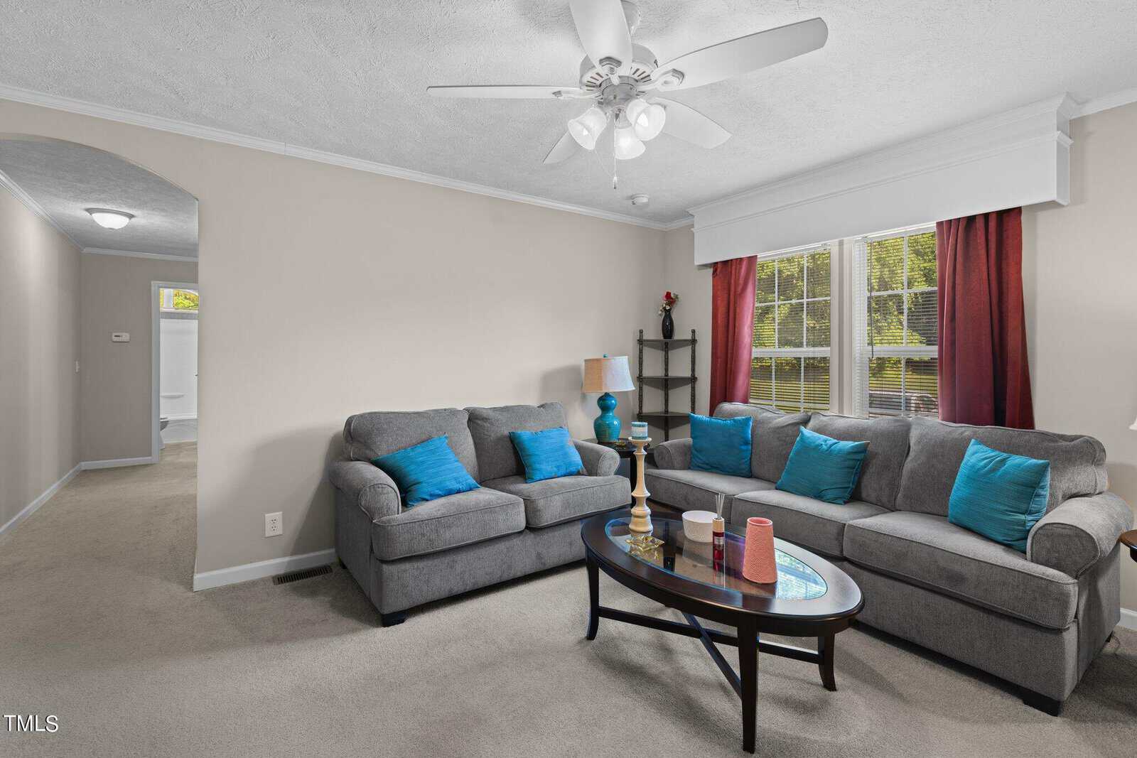 A cozy living room with gray sofas, blue cushions, and a dark wooden coffee table with candles. A ceiling fan and bright window with red curtains illuminate the room. A bookshelf is in the corner, and a hallway is visible in the background.