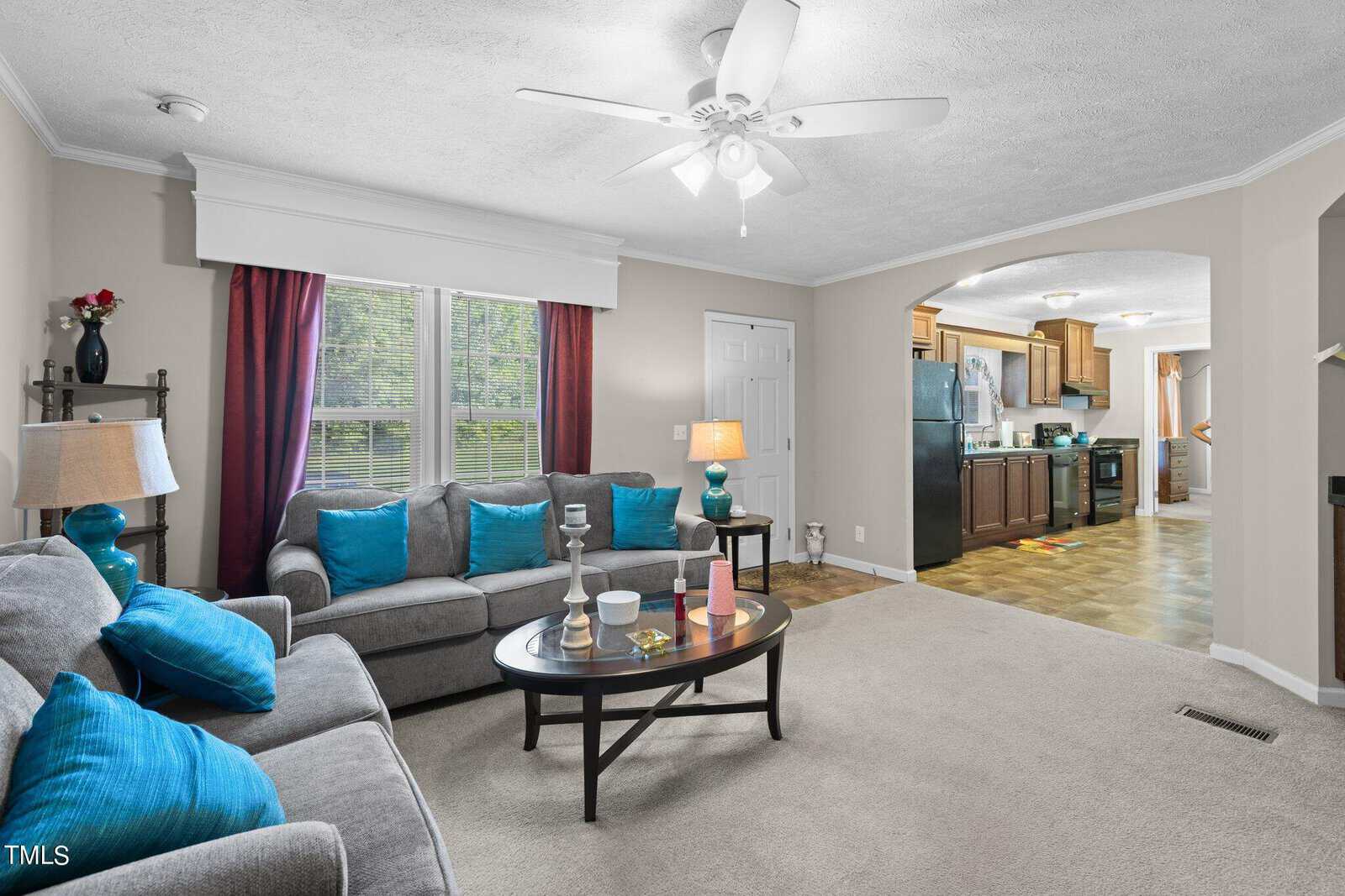 A cozy living room with gray sofas adorned with vibrant blue cushions. A black coffee table is centered on the beige carpet. In the background, a kitchen features wooden cabinets and stainless steel appliances. A ceiling fan is above.