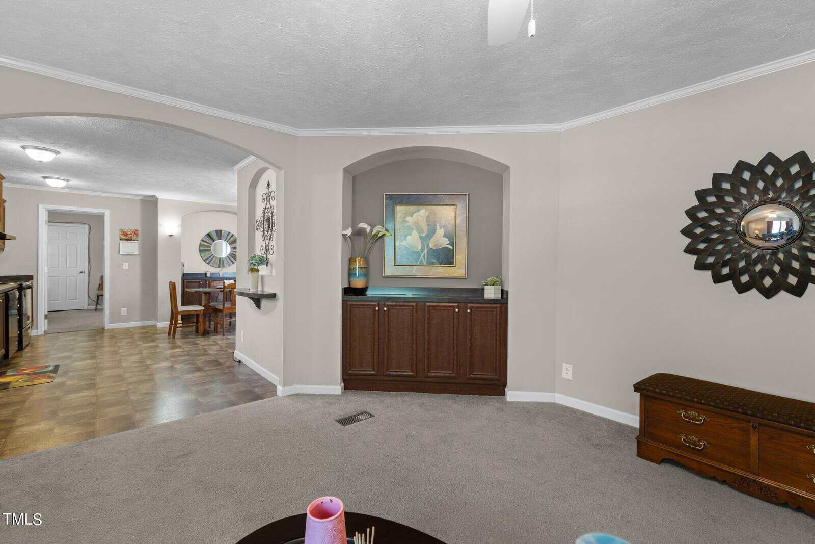 Modern living room with beige walls and a grey carpet. A dark wood cabinet with art pieces and plants sits in a recessed niche. The room features a decorative mirror and a view into a dining area with wood flooring.