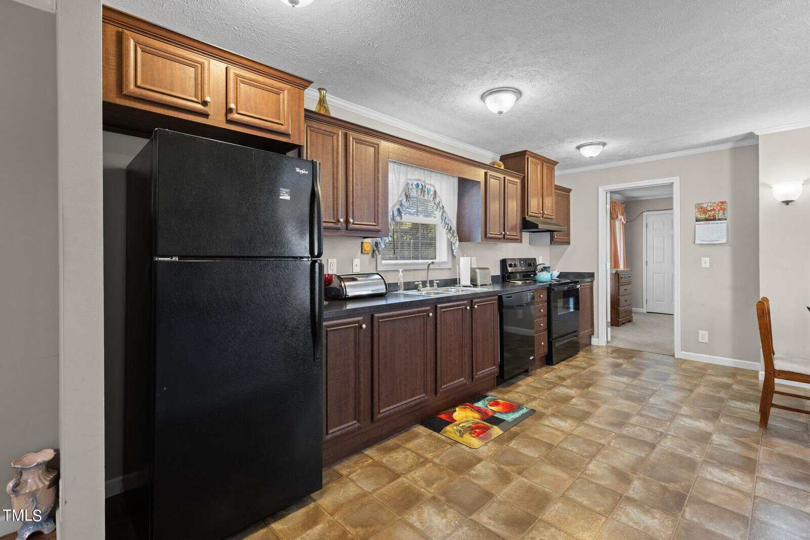 A kitchen with dark wooden cabinets, a black refrigerator, and a stove. There's a colorful rug on the tile floor and a doorway in the background. A framed picture hangs above the counter.