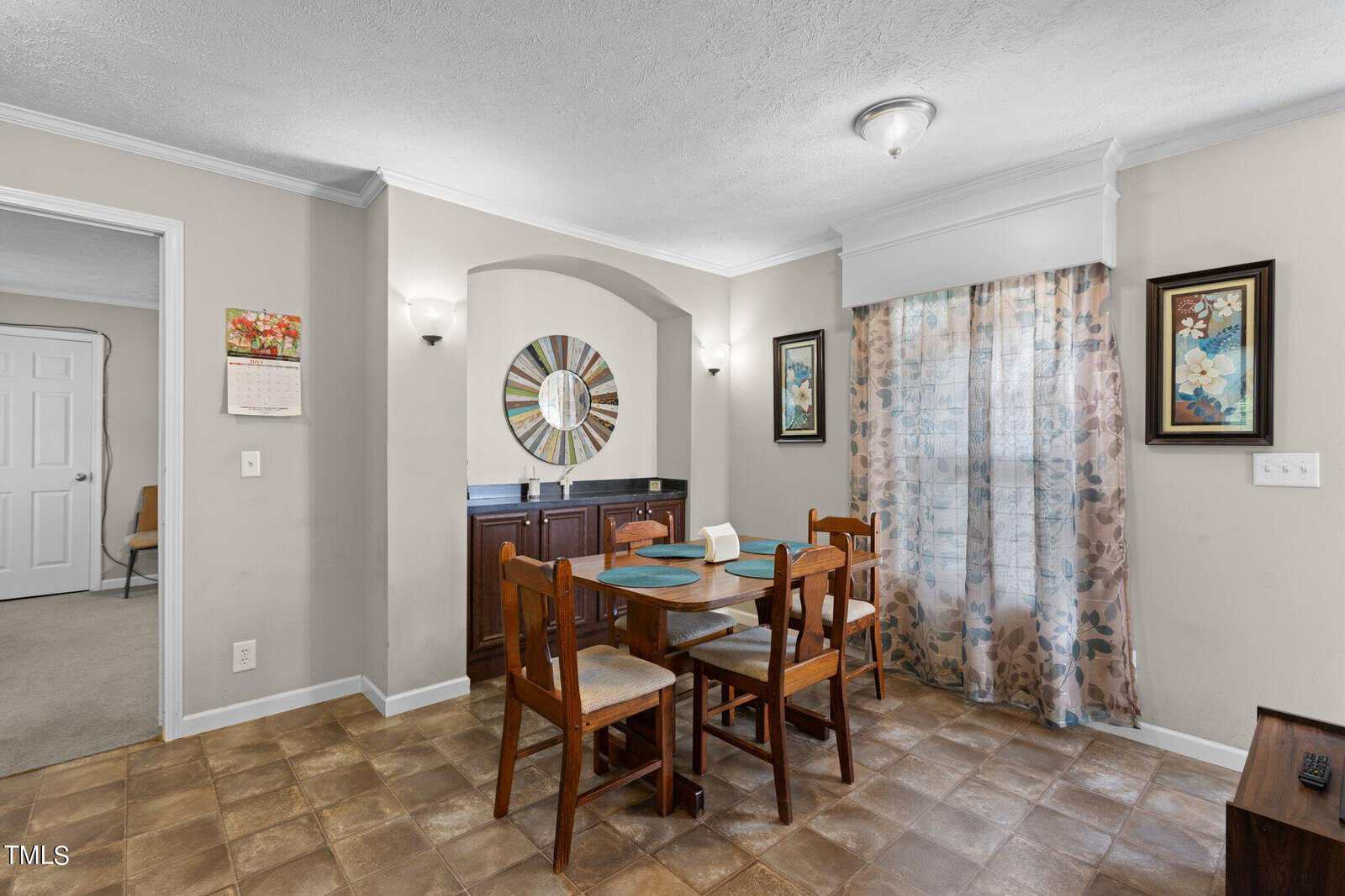 Dining area with a wooden table set for four, placed on tiled flooring. The walls are adorned with floral paintings and a round decorative mirror above a dark wood sideboard. A curtain covers a window, and a doorway leads to another room.