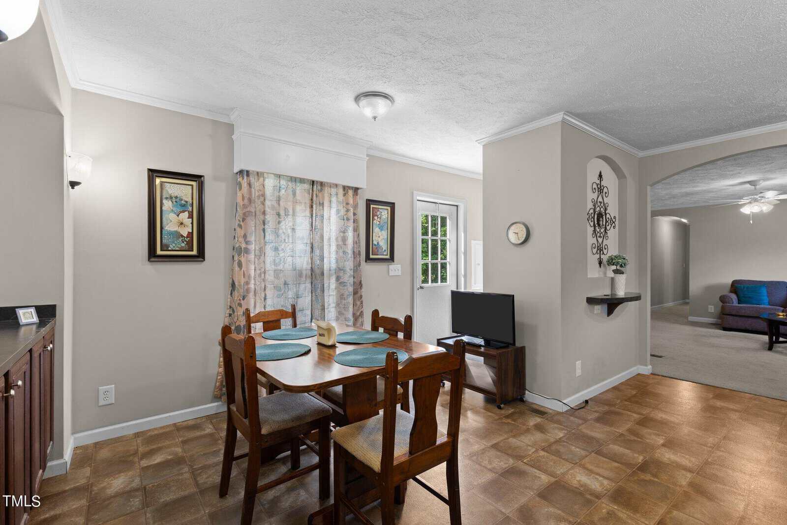 A cozy dining area with a wooden table set for four on a tiled floor. Adjacent is a small TV and a doorway with floral curtains. The walls are adorned with framed art, and a living space with sofas is visible in the background.