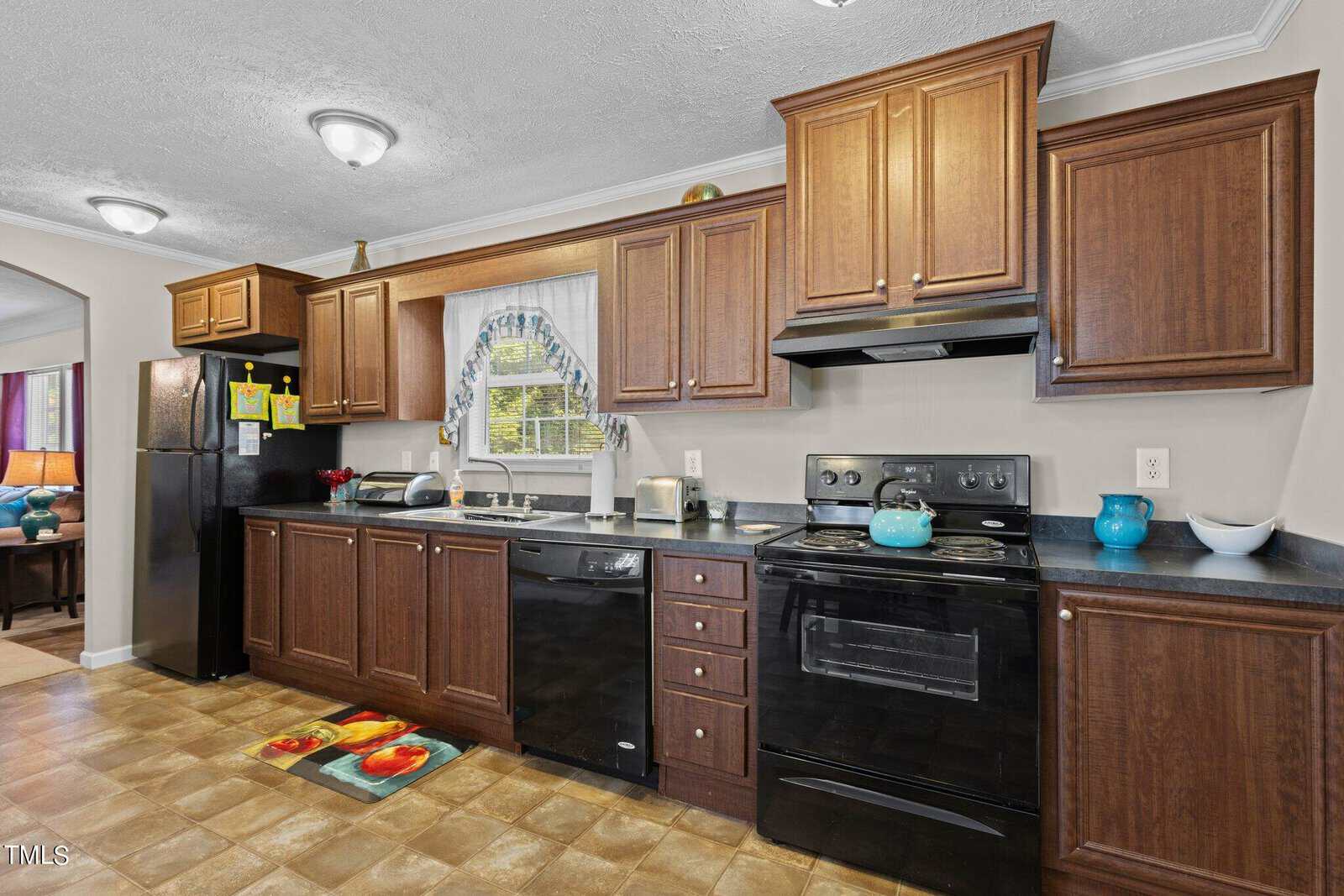 A kitchen with wooden cabinets, black appliances, and a black countertop. There is a stove, oven, fridge, and dishwasher. A small window with a floral curtain is above the sink. A colorful rug and overhead lights complete the look.