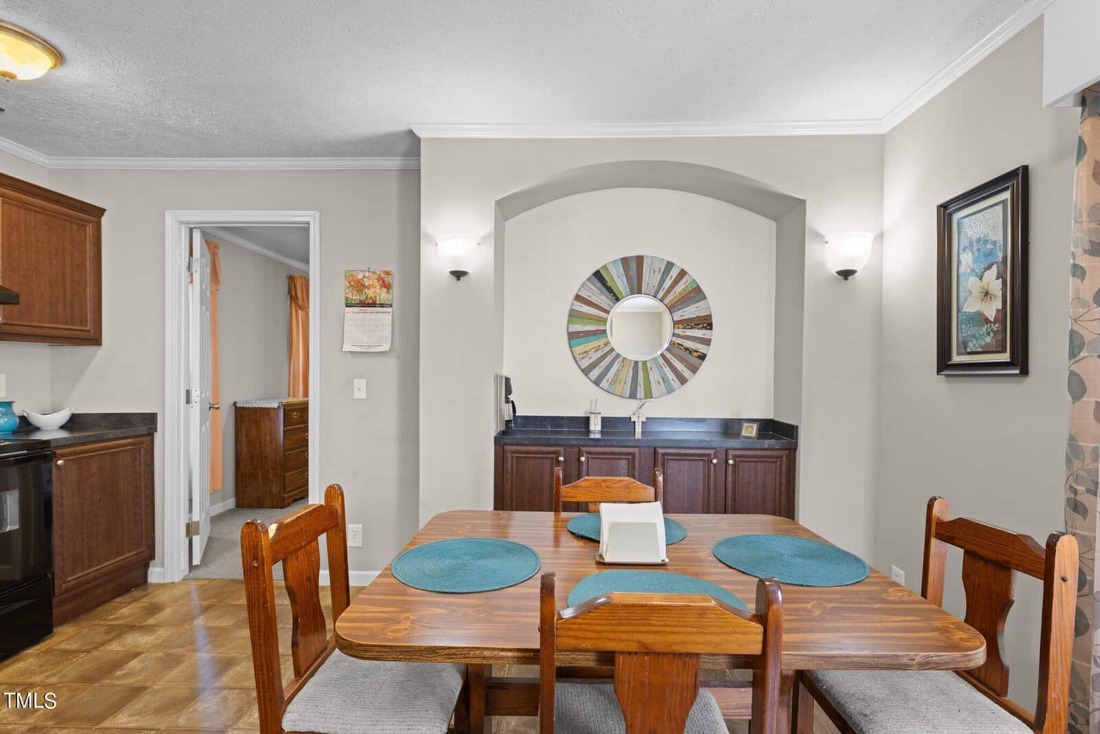 A dining room with a wooden table set for four, featuring teal placemats. The background has a wall alcove with a round mirror, surrounded by wall lights. Dark wooden cabinets are beneath the mirror, and a wall painting hangs on the right.