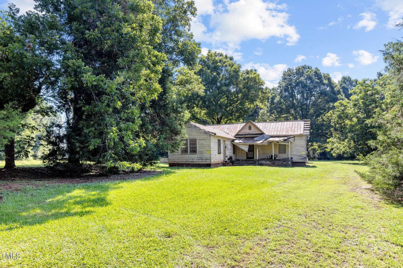 An old, weathered wooden house sits at 4764 Old Nc 75, surrounded by lush greenery and tall Oxford trees under a bright blue sky. The sunlit grassy lawn in front is wide, providing a tranquil rural setting.