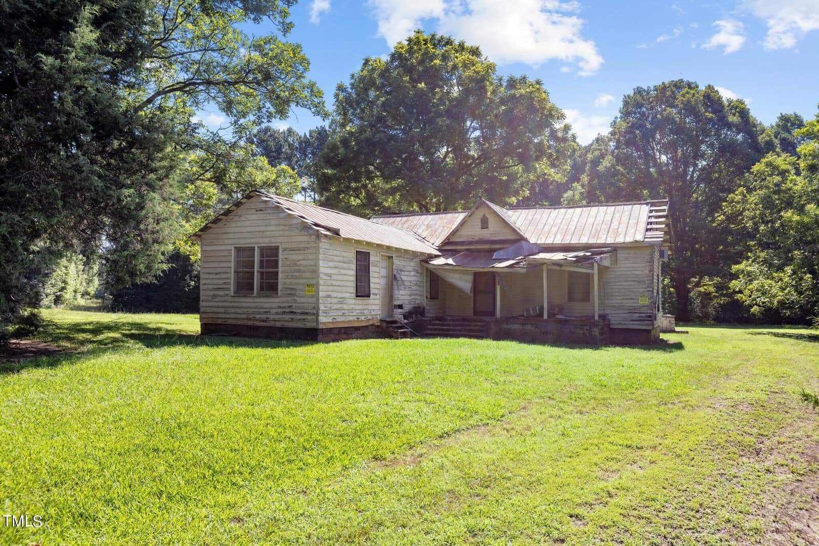 An old, abandoned wooden house with a rusty metal roof sits in a lush green field. Trees surround the property under a bright blue sky with scattered clouds. The house shows signs of wear and decay.