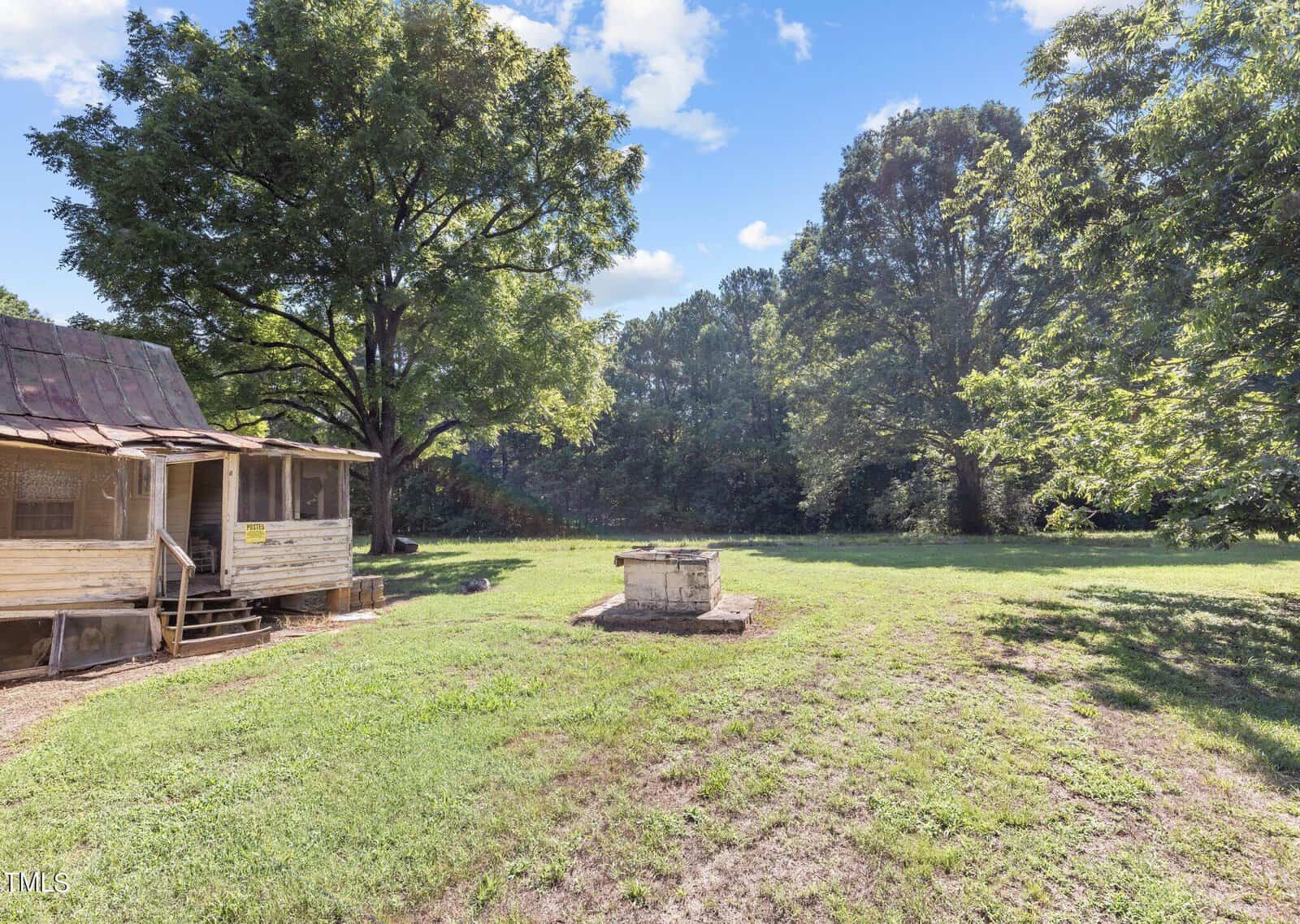 A rustic wooden cabin with a tin roof sits on a grassy yard, surrounded by tall trees. A stone well is in the center of the yard, under a clear blue sky with scattered clouds.