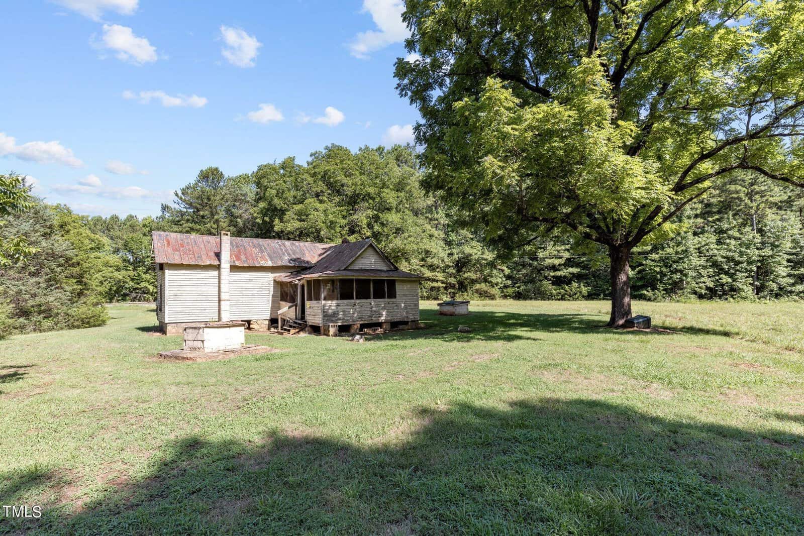 A small, rustic wooden cabin with a metal roof stands in a grassy, open area surrounded by trees. A large tree provides shade nearby, and a clear blue sky with a few clouds is in the background.