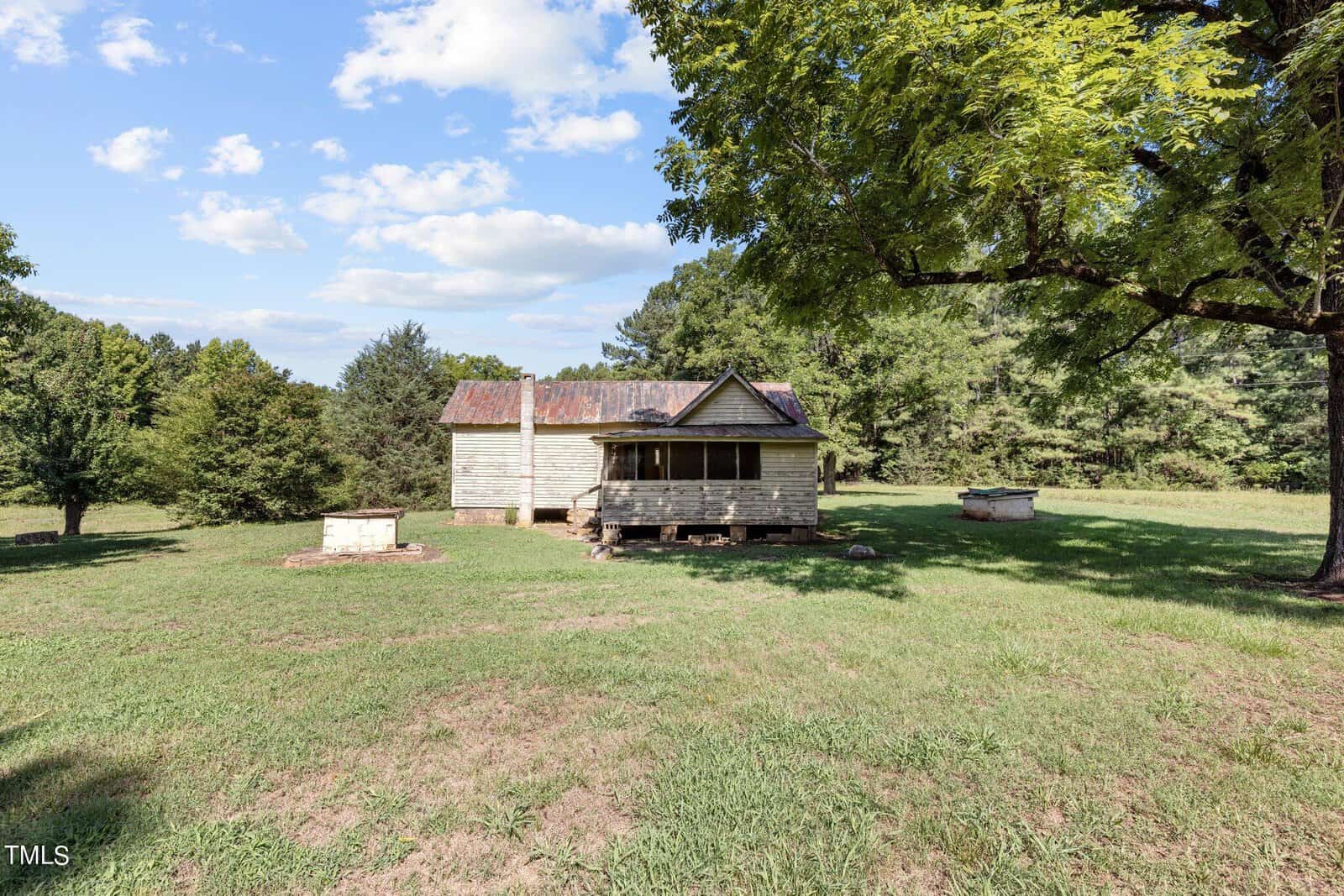 An old, weathered farmhouse with a rusty metal roof stands on a grassy plot, surrounded by trees under a partly cloudy sky. A large tree casts a shadow on the lawn, and a small well sits nearby.