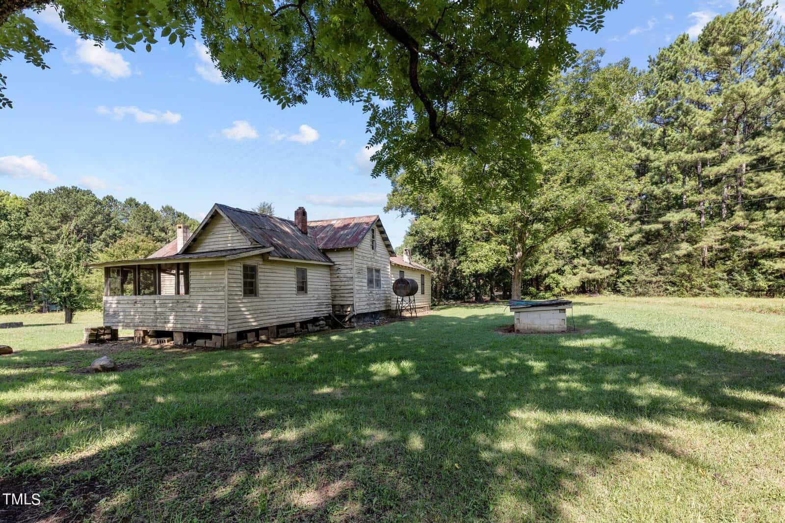 A rustic, wooden house with a metal roof sits in a grassy field surrounded by trees under a partly cloudy sky. There's a small, raised structure on the right and a large leafy tree providing shade in the foreground.