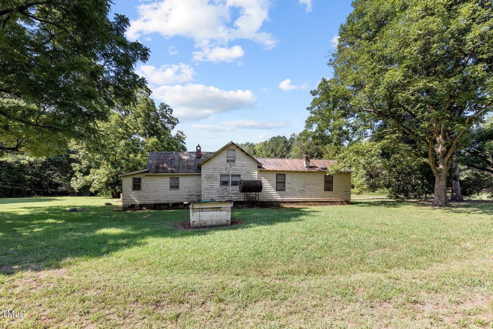 A weathered, single-story wooden house with a rusting roof stands in a grassy field. Trees surround the property under a partly cloudy sky. A small wooden structure is in the foreground.
