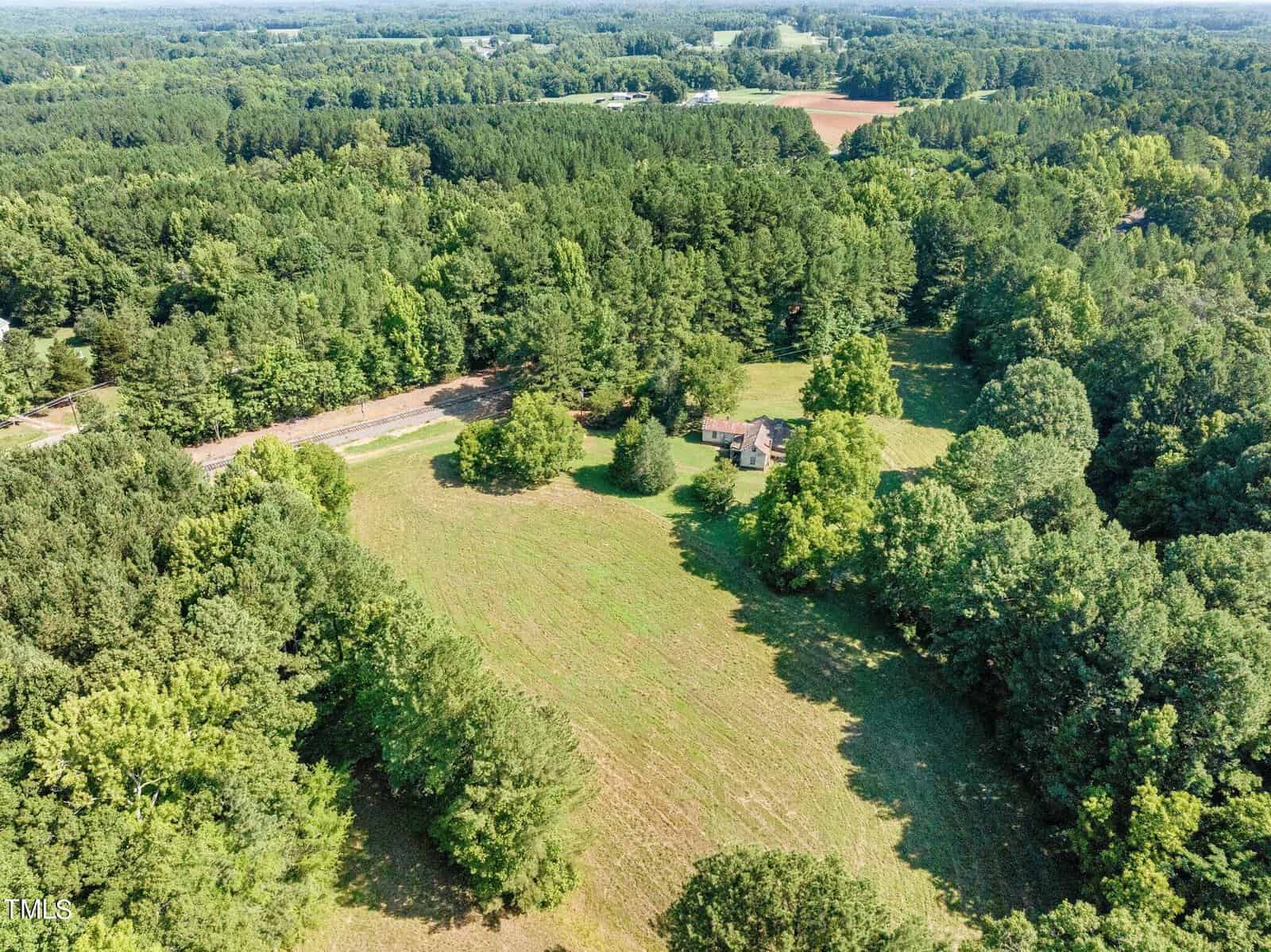 Aerial view of a rural landscape featuring a large green field surrounded by dense forest. In the middle of the field is a small house. Roads are visible in the distance, and the horizon shows expansive wooded areas.