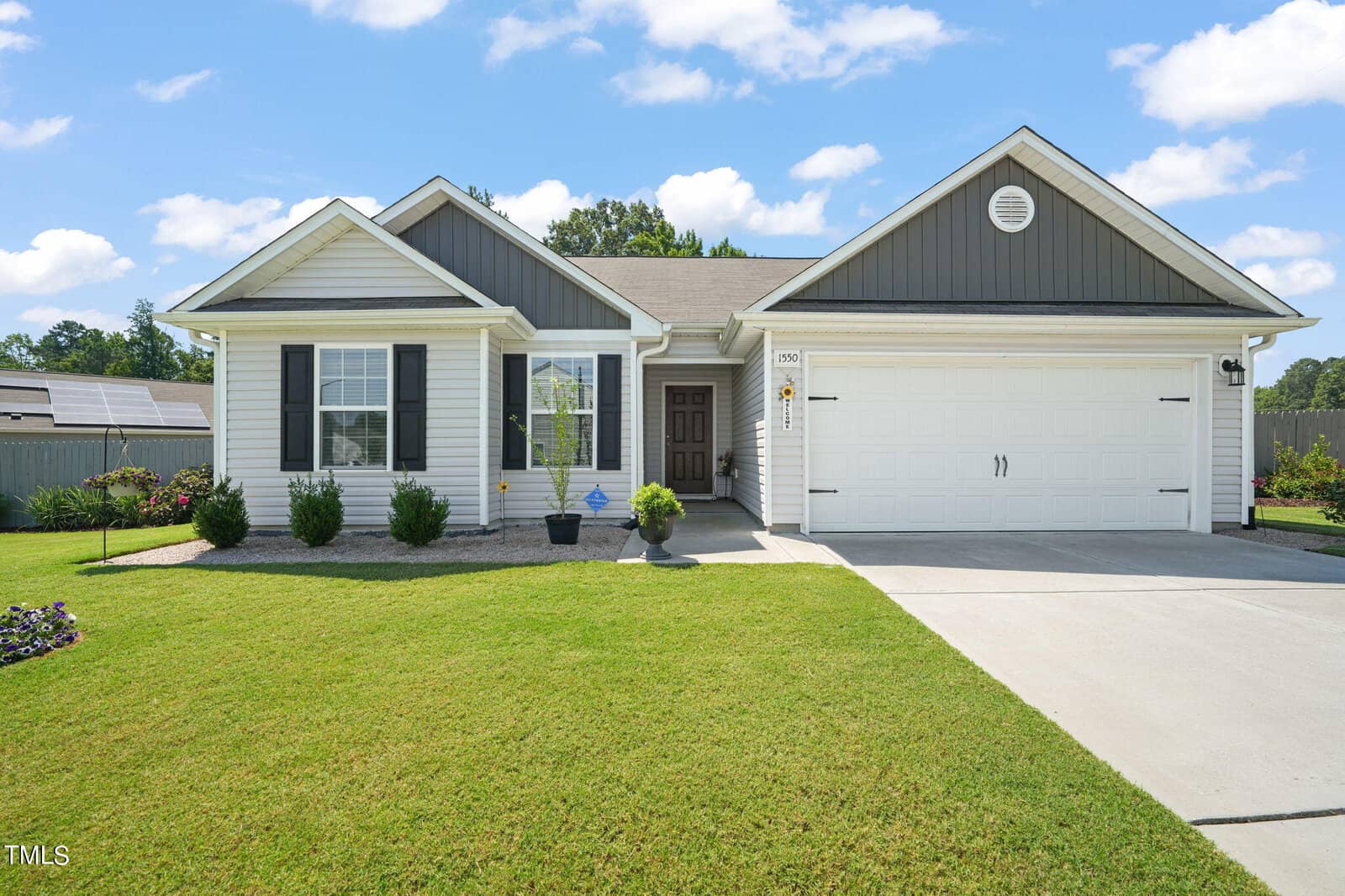 Located at 1550 Alderman Way in Creedmoor, this charming suburban one-story house features a gray roof and white siding. It boasts a two-car garage and a cozy porch with a potted plant, all surrounded by a well-maintained lawn under a blue sky with scattered clouds.