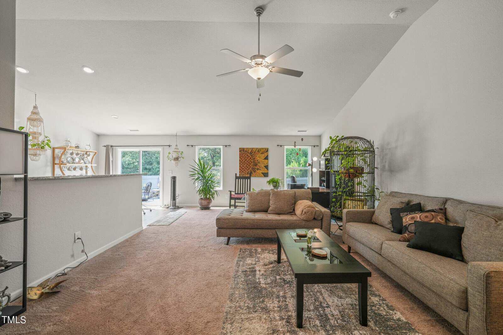 Cozy living room with beige sofas, a black coffee table, and a ceiling fan. Large windows let in natural light. Walls are decorated with plants and artwork. A birdcage is visible, and the dining area is in the background.