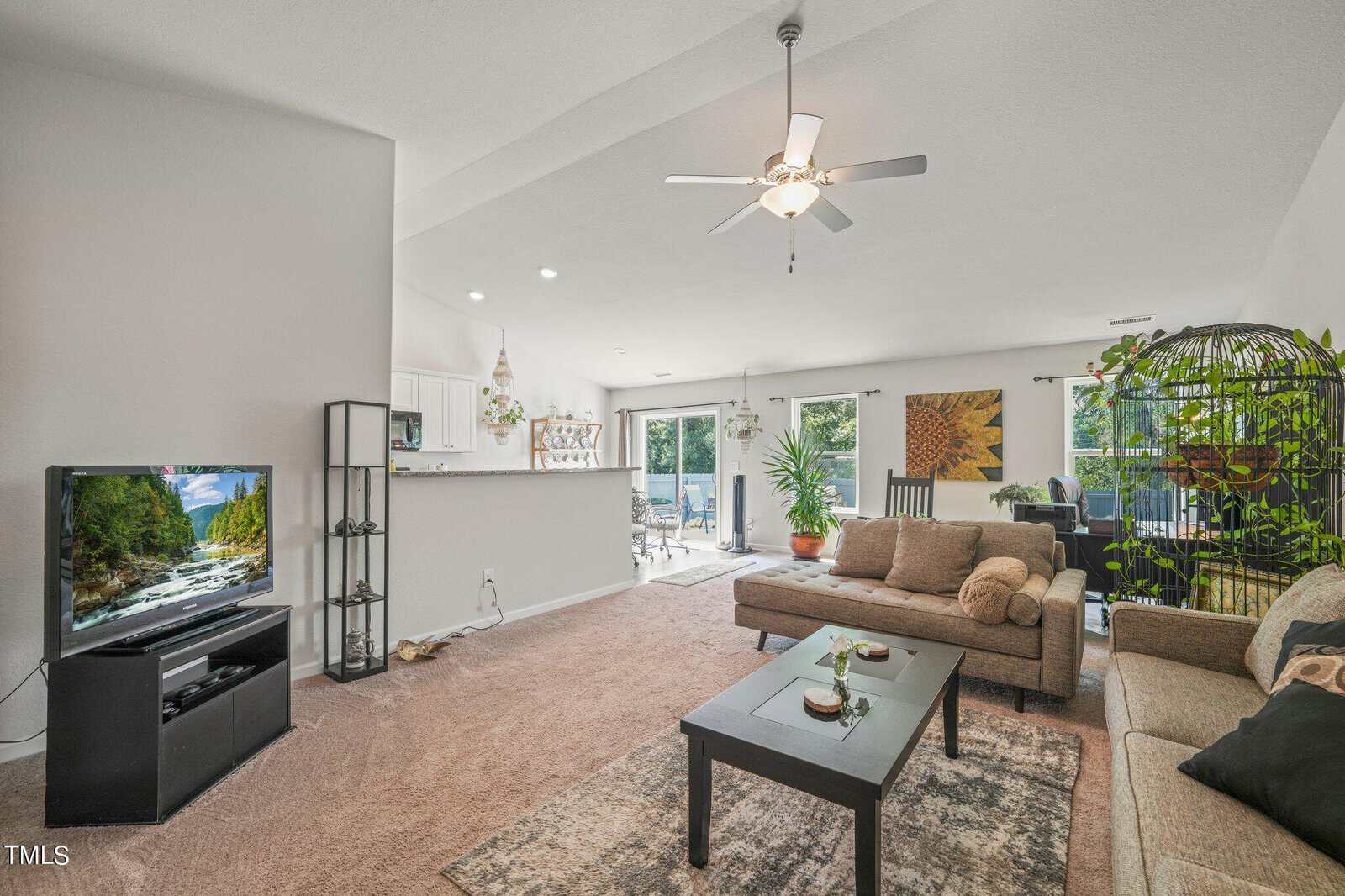 Spacious living room with beige carpet, a ceiling fan, and neutral-colored sofas. There's a dark coffee table, large TV, decorative display shelf, and a birdcage. In the background, a dining area with windows and light streaming in.
