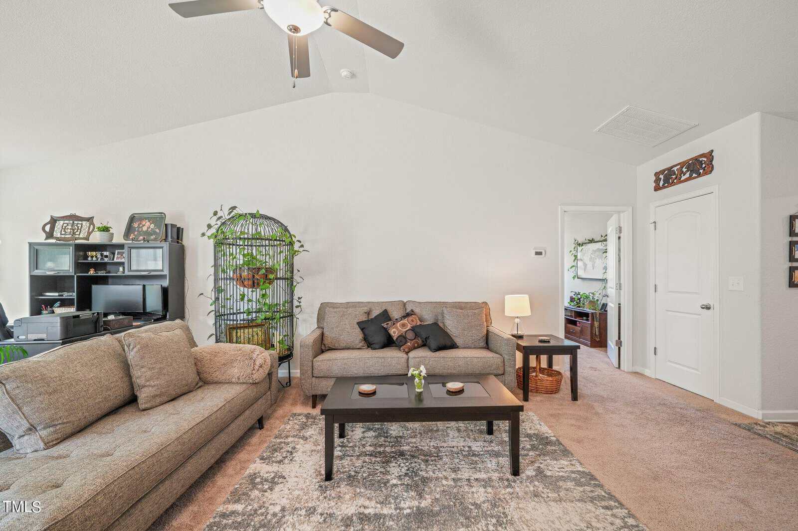 A cozy living room with two beige sofas, a black coffee table, and a patterned rug. A tall plant and decorative items occupy the space. A ceiling fan is overhead, and a glimpse of an adjacent room is visible through an open door.