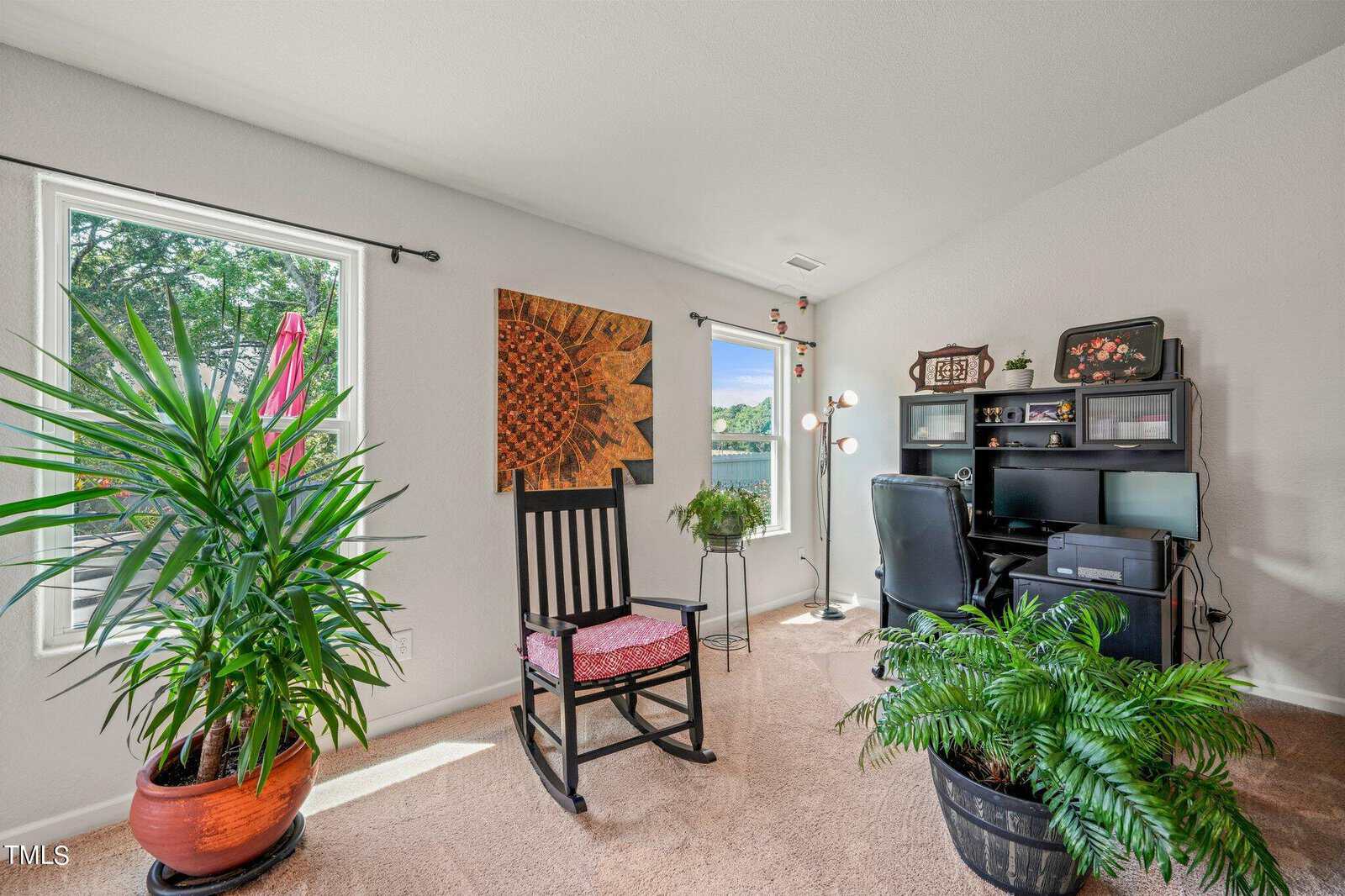 A bright home office with large windows featuring a rocking chair, plants in pots, a computer desk with a black office chair, and wall art. Light streams in from outside, illuminating the cozy and inviting space.