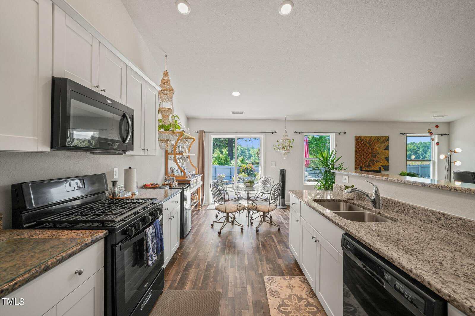 Modern kitchen with dark wood flooring, granite countertops, and white cabinets. A stove and microwave are on the left, and a double sink is on the right. Dining area with a round table and chairs near large windows showing a view outside.