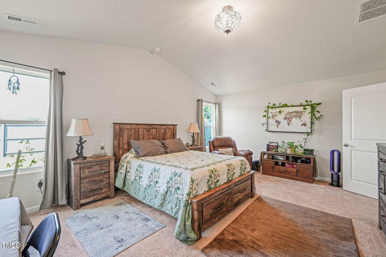 A cozy bedroom featuring a wooden bed with a floral green and beige bedspread, matching nightstands with lamps, and a brown armchair. A wall map decorates the room, which has carpeted floors and large windows for natural light.