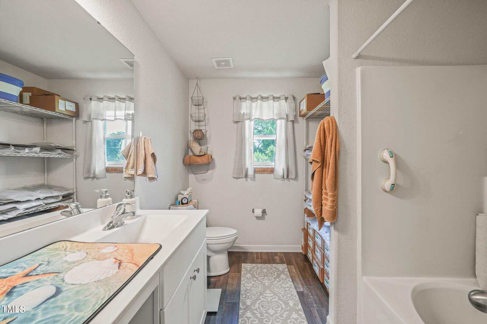 A small bathroom featuring a double sink with a seashell-themed mat, a toilet, and a shower with a grab bar. Shelves hold towels and boxes. Two windows with valances brighten the room, while a rug adds warmth to the wooden floor.