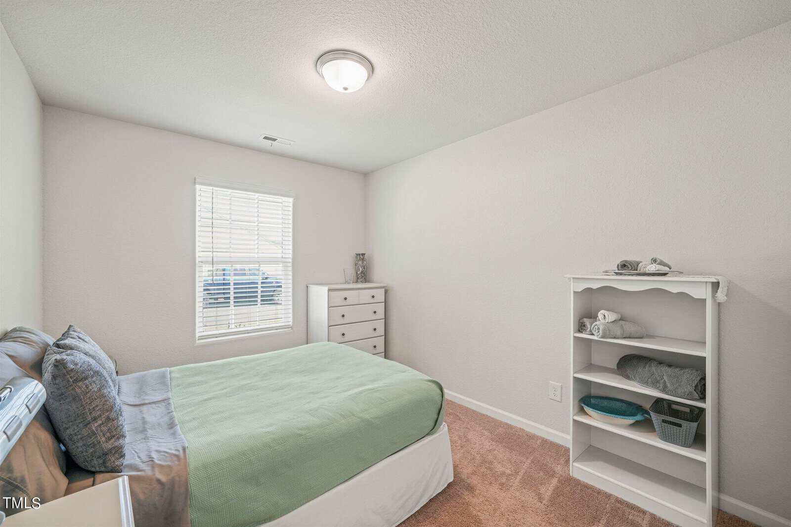 A small bedroom with a neatly made bed featuring a green comforter and gray pillows. There is a white dresser with a vase on top by the window and a white shelving unit with folded towels and baskets on the right. The room has beige carpet and a ceiling light.