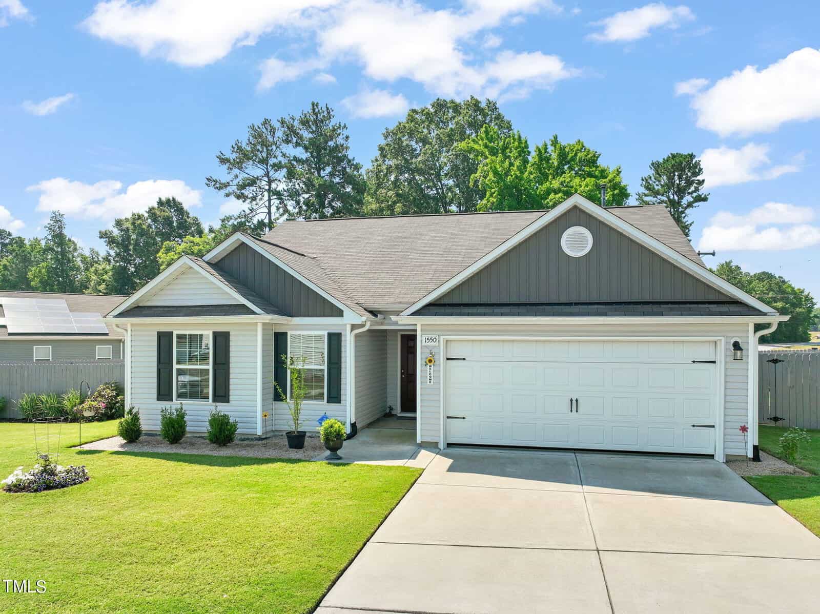 Single-story house with white siding and dark gray accents. It has a neatly maintained yard, two-car garage, and a concrete driveway. Bushes are planted along the front, with a clear blue sky and trees in the background.