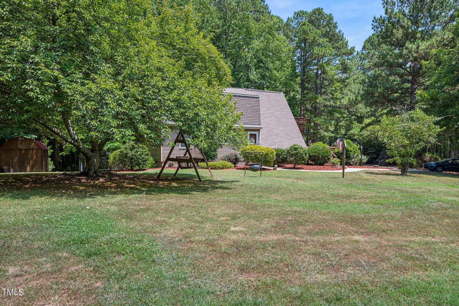 A house with sloped roofs partially hidden behind trees, surrounded by a large, grassy yard. A wooden swing set stands in the foreground under a leafy tree. The yard is bordered by a dense line of trees. A car is parked on the right side beyond the house.