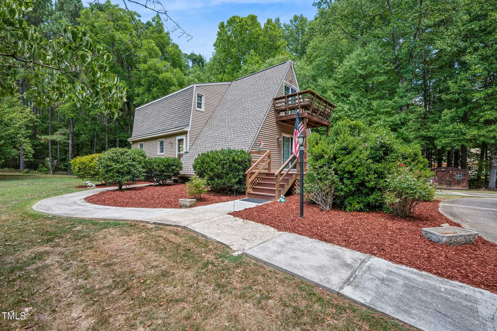 Located at 1817 Ruin Creek Road in Henderson, NC, this house boasts a unique triangular roof design and features a wooden staircase leading up to a balcony with a railing. Surrounded by greenery, including bushes and large trees, the home also has a manicured lawn and a red mulch path.