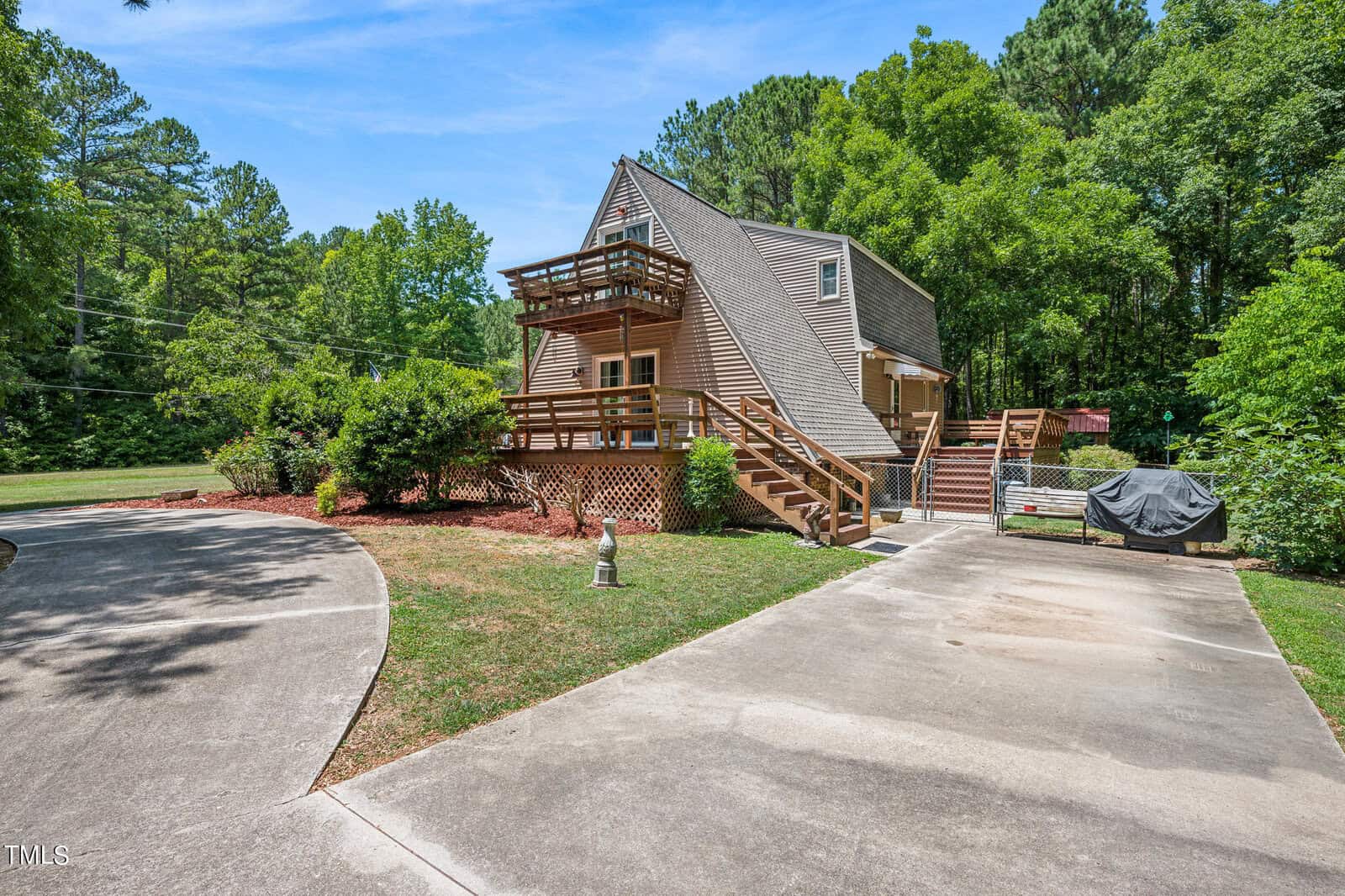 A unique, triangular-shaped house with multiple decks surrounded by lush greenery. The driveway is curved, leading to the entrance of the house. There is an outdoor grill near the house and a small garden area adjacent to the driveway.