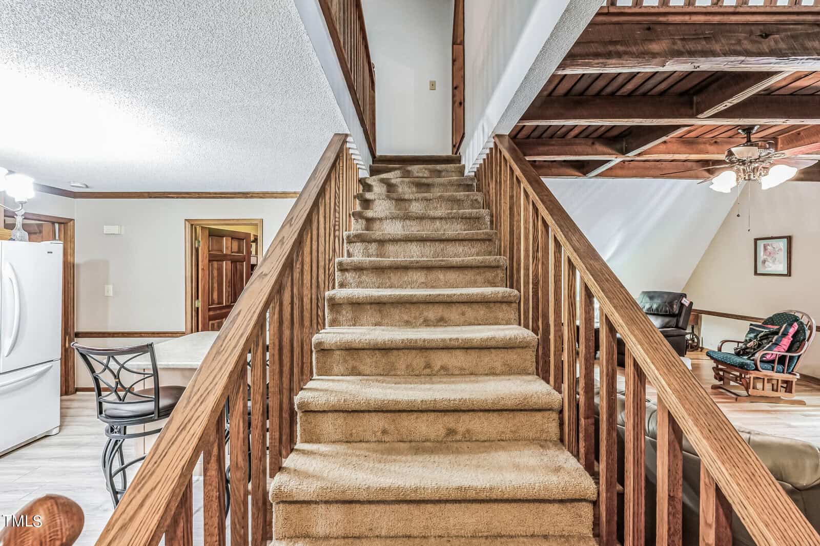 A rustic interior features a carpeted staircase with wooden railings leading to an upper loft. To the left, a kitchen with white appliances and a barstool is visible. To the right, a living area is partially shown with a ceiling fan, armchairs, and artwork on the wall.