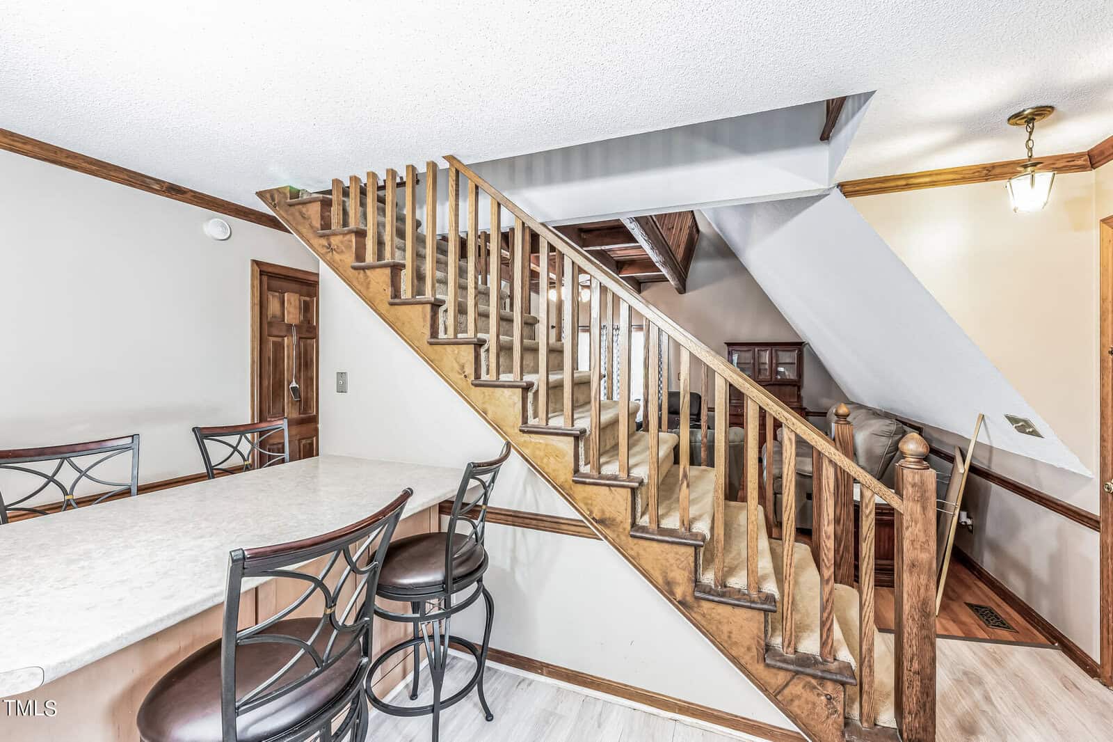 A cozy interior features a wooden staircase with carpeting, leading up to a second level. Adjacent to the staircase is a small breakfast bar with white countertop and dark metal barstools. Warm wood trim accents the walls and the ceiling.